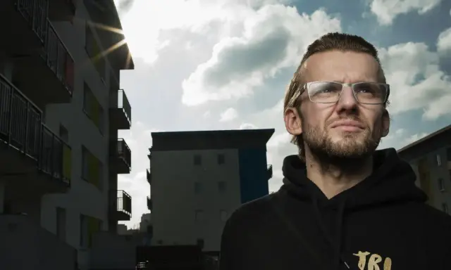 Mężczyzna w okularach patrzy w górę, na tle bloków mieszkalnych. Słońce przebija się przez chmury, tworząc efekt kękę dzieci.