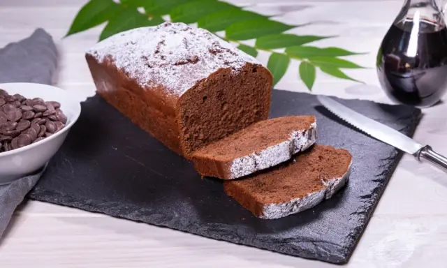 Ein saftiger Schokoladenkuchen, bestäubt mit Puderzucker, bereit für ein köstliches rotweinkuchen rezept. Daneben Schokoladendrops und ein Glas Rotwein.