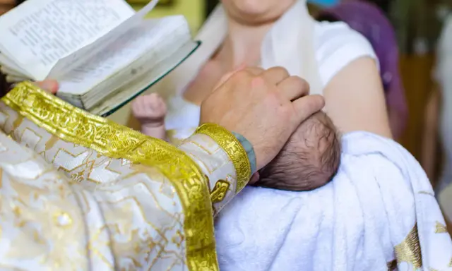 Ksiądz w złotej szacie trzyma dziecko do chrztu, czytając z księgi.