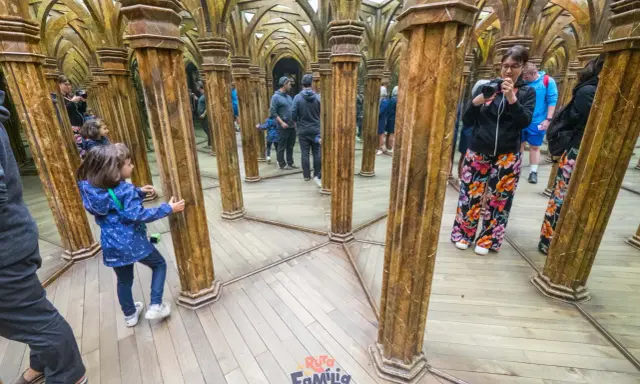 Niños exploran un laberinto de espejos con columnas doradas, creando un divertido juego de "praga con niños" y reflejos infinitos.