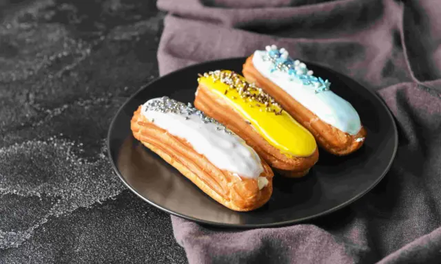 Trois éclairs gourmands sur une assiette noire : un blanc pailleté, un jaune croquant, un bleu ciel festif. La pâte à choux éclair inratable est prête à être dégustée.