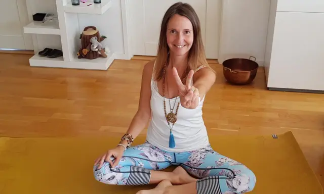 En kvinna sitter i lotusställning på en gul yogamatta, redo för morgon meditation. Hon ler och gör en fredstecken.