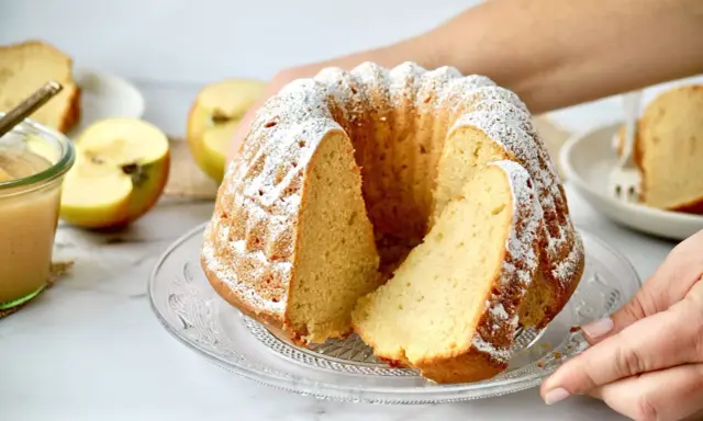 Ein saftiger Kuchen mit Apfelmus, bestäubt mit Puderzucker. Daneben liegen Äpfel und ein Glas Apfelmus.