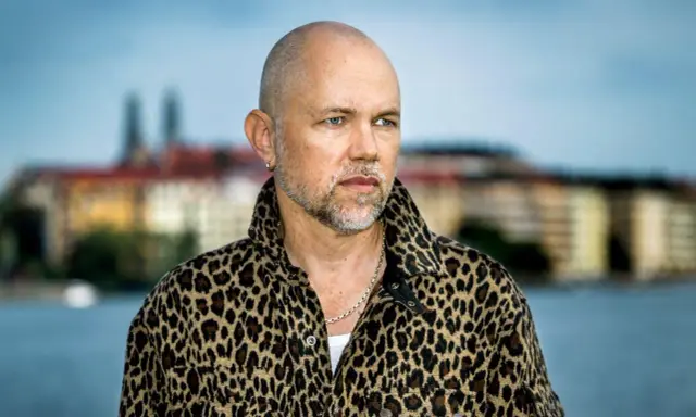 Peter LeMarc i leopardmönstrad jacka, redo för en konsert. Bakgrunden visar en stad vid vattnet.