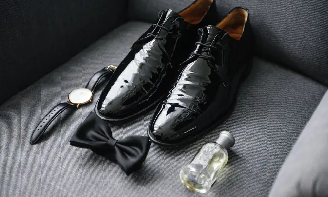 Shiny black patent leather shoes to wear with tuxedo, a bow tie, watch, and cologne are laid out on a grey couch.
