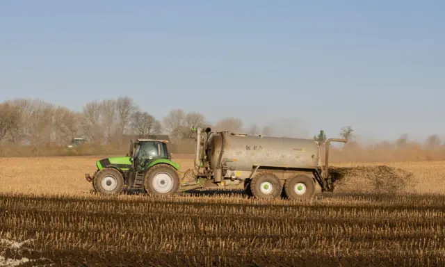 Zielony traktor z przyczepą rozprowadza obornik bydlęcy na polu.