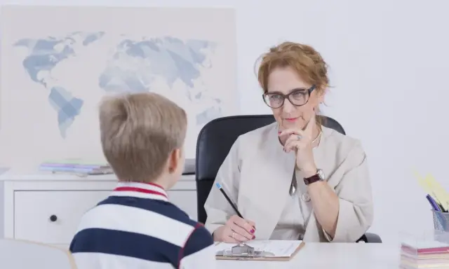 Chłopiec rozmawia z nauczycielką o ocenach. Nauczycielka słucha uważnie, notując coś na kartce.
