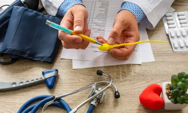 Lekarz analizuje formularz medyczny, trzymając model plemnika i długopis. Na stole leżą stetoskop, ciśnieniomierz, młoteczek neurologiczny i leki, sugerując dyskusję o skutkach ubocznych wazektomii.