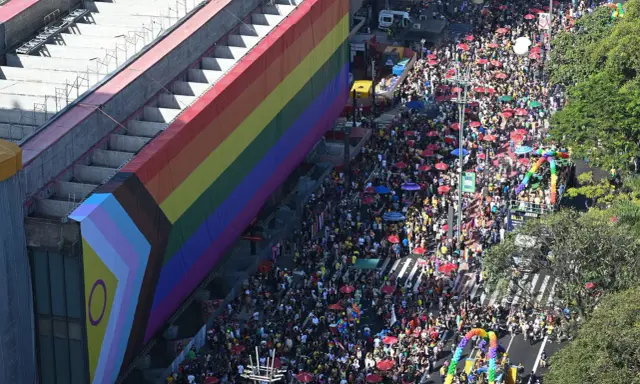 A massive crowd fills the street for what appears to be the biggest pride parade, with a giant rainbow flag draped over a building.