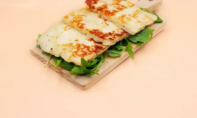Trzy grillowane plastry sera halloumi, z czego jest, ułożone na desce z rukolą.