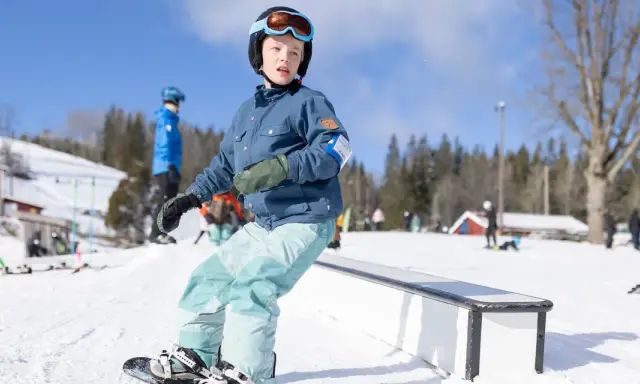 Ungdom åker snowboard i backen, redo för nästa trick. Solen skiner och snön ligger vit.