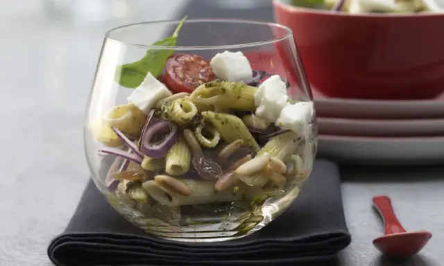 Ein köstlicher Nudel Pesto Salat mit Mozzarella, Pinienkernen, Rosinen und roten Zwiebeln in einem Glas serviert.