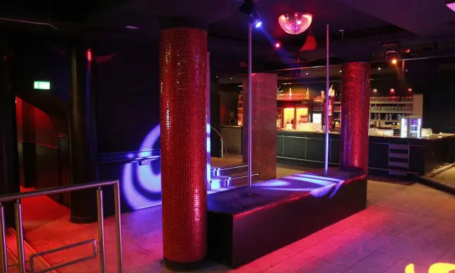 Interno di una boite de nuit con luci soffuse, un palo da lap dance, un bancone bar e una sfera da discoteca.