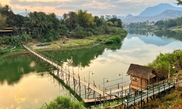 Drewniany mostek nad rzeką, bambusowa chatka i góry w tle. To jedne z wielu **laos atrakcje** warte zobaczenia.