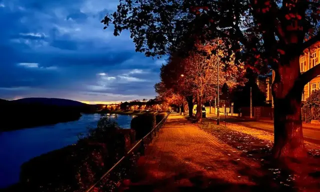Un sentiero illuminato da lampioni lungo un fiume, con alberi dalle foglie arancioni e rosse. Una magica sera d'ottobre sui pascoli.