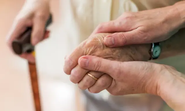Dwie dłonie, jedna starsza, druga młodsza, trzymają się nawzajem. Laska w tle sugeruje trudności z poruszaniem się, być może związane z chorobą Parkinsona i jej dziedziczeniem.