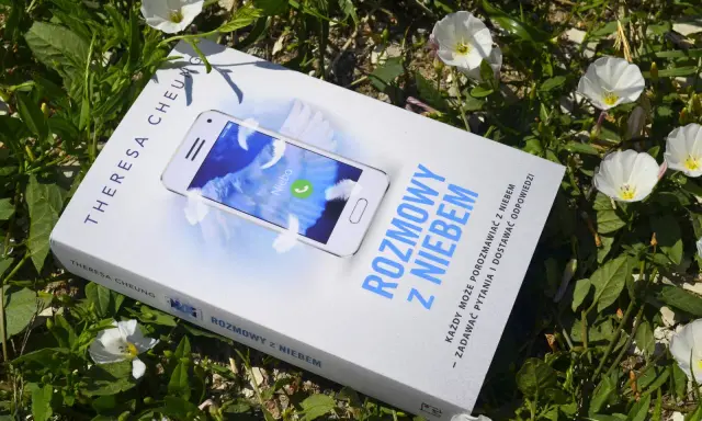 Książka "Rozmowy z niebem" leży wśród zieleni i białych kwiatów. Na okładce telefon z napisem "Niebo" i piórami.