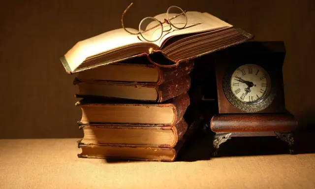 Un libro aperto con occhiali da lettura, una pila di antichi volumi e un orologio da tavolo. Un invito a leggere Catullo: "dammi mille baci".