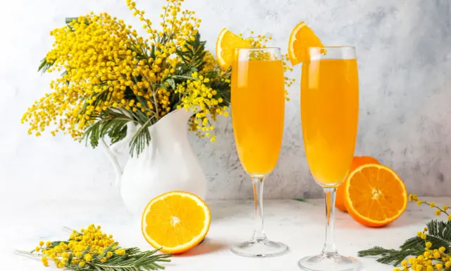 Dwa kieliszki orzeźwiającego drinka mimosa, ozdobione plasterkami pomarańczy, stoją obok bukietu żółtych kwiatów akacji.