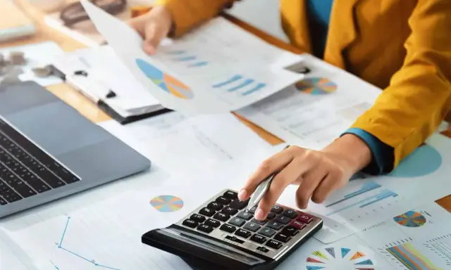 Persoon berekent cijfers op een calculator, omringd door grafieken en documenten. Dit kan te maken hebben met de commissie sociaal minimum.