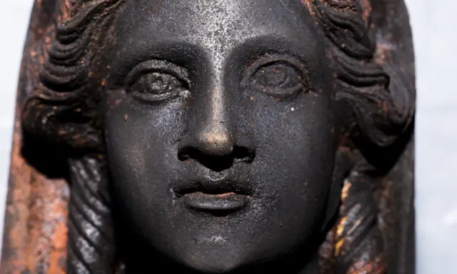 Volto di bronzo scuro, un dettaglio dei bronzi di San Casciano dei Bagni, con capelli a onde e trecce.