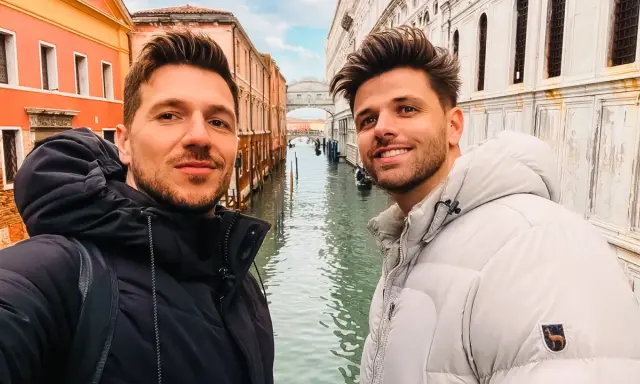 Two men enjoying a romantic gay Venice trip, smiling for a selfie with canals and historic buildings behind them.