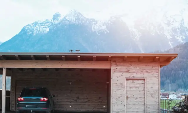 En nybyggd carport i trä med en bil parkerad under. Bakom syns snöklädda berg.
