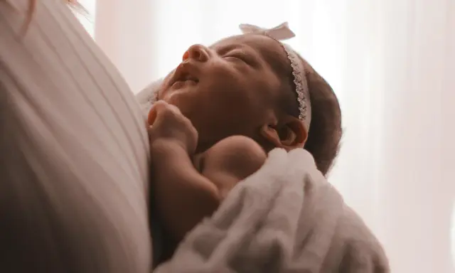 Un neonato avvolto in una coperta bianca, con un fiocco rosa in testa. I suoi piccoli tremori sono quasi impercettibili mentre dorme serenamente.