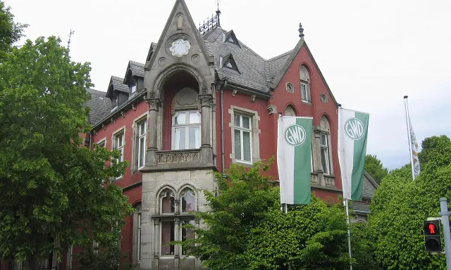 Die Kaiservilla in Minden, ein rotes Backsteingebäude mit Türmchen und zwei GWD-Fahnen, umgeben von üppigem Grün.