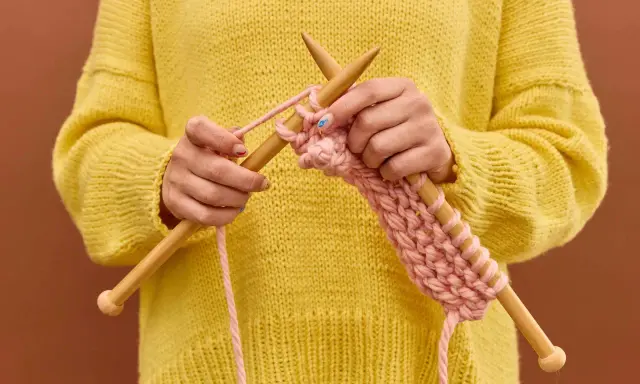 Manos tejiendo un suéter amarillo con hilo rosa, mostrando cómo hacer ochos de 4 puntos.