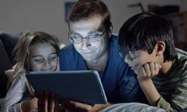 Padre y dos hijos ven una tablet, sonriendo. Podrían estar aprendiendo sobre los riesgos de las redes sociales o disfrutando de contenido familiar.