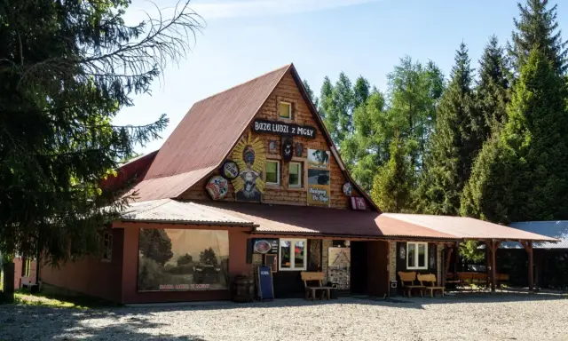 Drewniany budynek z napisem "Baza Ludzi z Mgły" i malowidłami. Obok duży mural z krajobrazem i napisem "Baza Ludzi z Mgły".