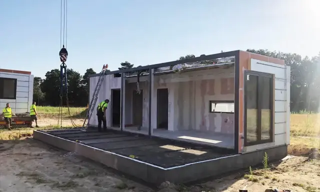 Budowa domu na zgłoszenie postępuje. Dźwig podnosi moduł, a pracownicy w kamizelkach nadzorują montaż. Warunki zabudowy sprzyjają szybkiemu postawieniu konstrukcji.