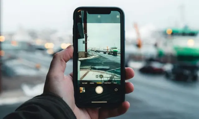 Dłoń trzyma aparat na telefon, rejestrując zimowy widok portu z zielonym statkiem i samochodami na drodze.