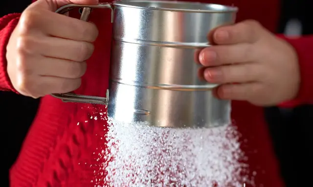 Hände halten einen Siebbecher, aus dem feiner Zucker rieselt. So einfach lässt sich puderzucker selber machen für süße Kreationen.