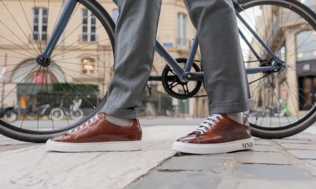Eleganckie sneakersy do garnituru w brązowym kolorze, zestawione z szarymi spodniami, na tle miejskiego krajobrazu z rowerem.