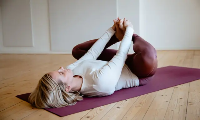 Woman in white top and burgundy leggings lying on a purple yoga mat, holding her feet.