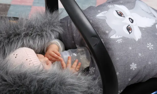 Noworodek w kombinezonie z futerkiem i czapką trzyma butelkę z mlekiem w foteliku samochodowym.