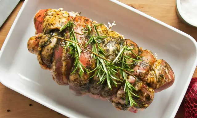 Un rôti de porc cru, assaisonné et garni de romarin frais, prêt à être cuit dans un plat blanc.