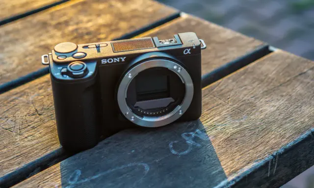 Czarny aparat Sony Alpha ZV-E10 bez obiektywu leży na drewnianym stole, oświetlony ciepłym światłem zachodzącego słońca.