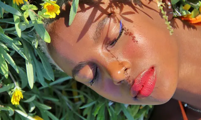 Un volto immerso tra fiori, con un trucco festival audace: eyeliner blu elettrico e labbra rosse vibranti.