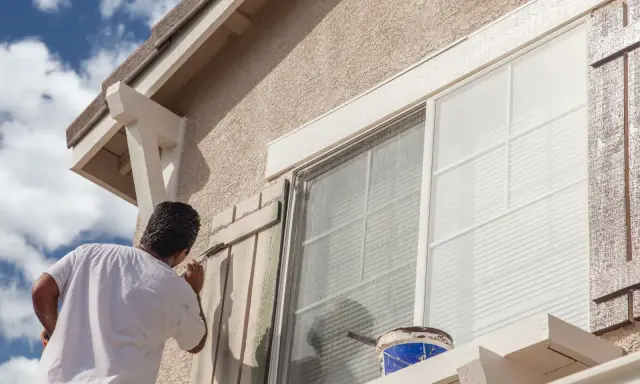 Un hombre pinta las contraventanas de una casa. El trabajo de pintar casas por fuera es esencial para su mantenimiento.