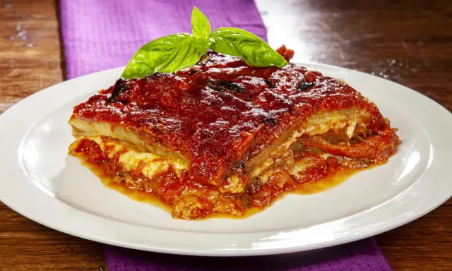 Una fetta di parmigiana di melanzane al forno, ricca e succulenta, guarnita con basilico fresco.