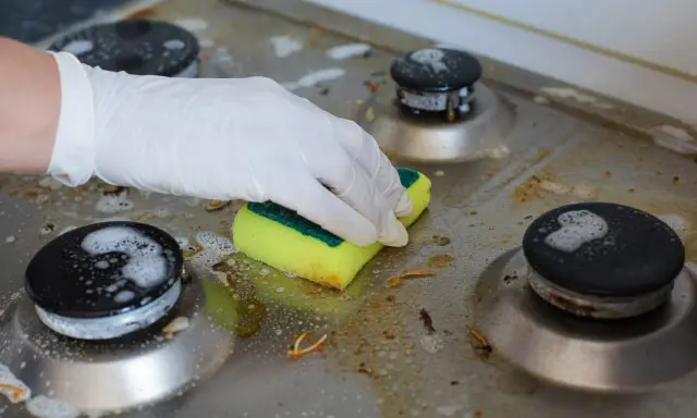 Ręka w rękawiczce czyści zabrudzoną kuchenkę gazową gąbką. Poznaj sposoby, jak wyczyścić kuchenkę, by znów lśniła.