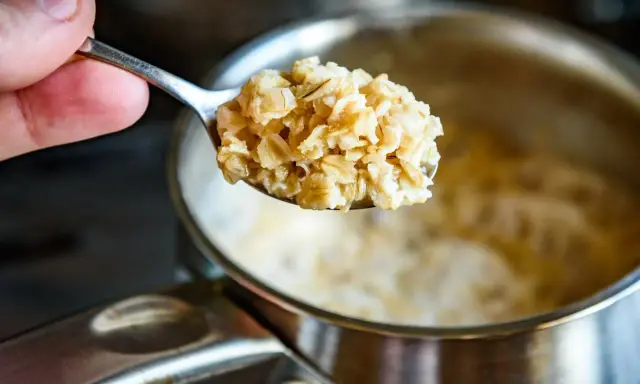 Łyżka gotowanych płatków owsianych, które pomagają obniżyć cholesterol. W tle garnek z owsianką.