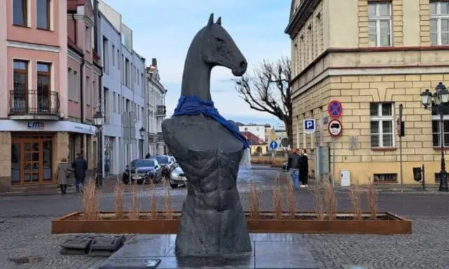 Pomnik konia w Koninie, z niebieską szarfą na szyi, stoi na brukowanym placu, otoczony kamienicami.