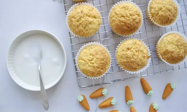 Goldbraune Karottenmuffins auf einem Gitter, daneben eine Schüssel mit Zuckerguss und süße Marzipankarotten. Perfekt für ein Karottenmuffins Rezept!
