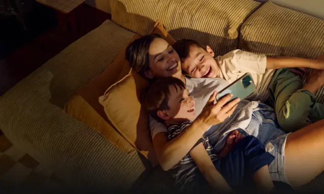 A mother and two sons laugh while looking at a smartphone on a couch.