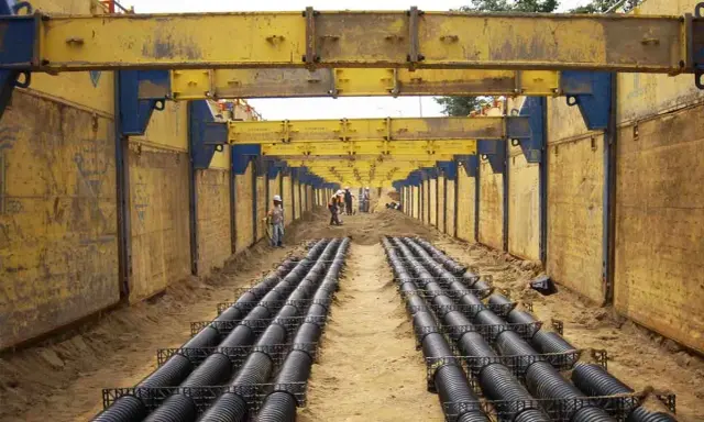Trabajadores instalan tuberías en una zanja profunda, con estructuras metálicas amarillas para entibar zanjas y prevenir derrumbes.