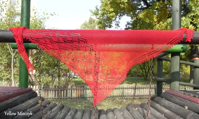 Czerwony, ażurowy szal, wykonany według darmowych wzorów z Ravelry, wisi na drewnianej konstrukcji placu zabaw.